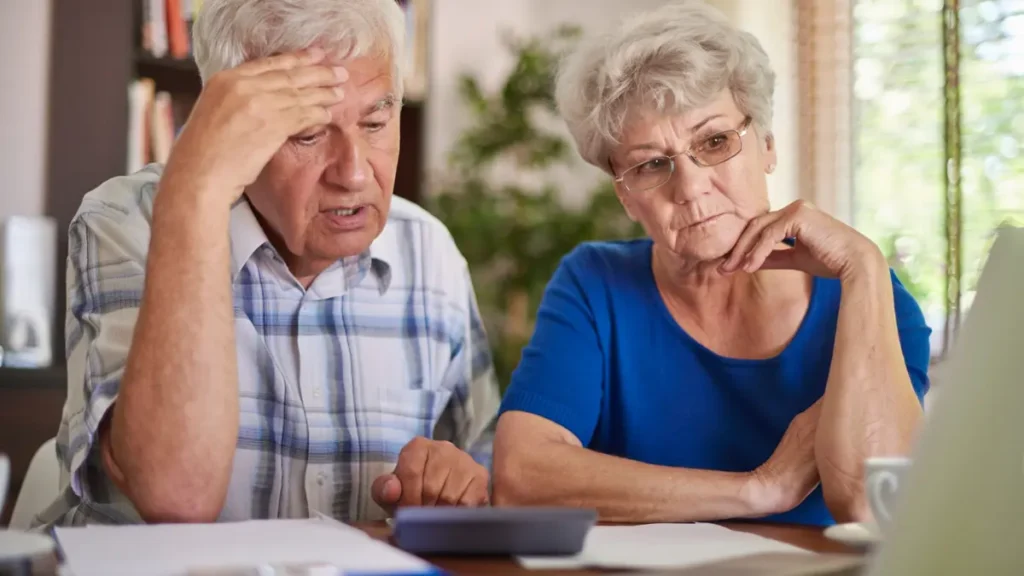 Elderly couple looking stressed while reviewing bills and financial paperwork together at home with a calculator.