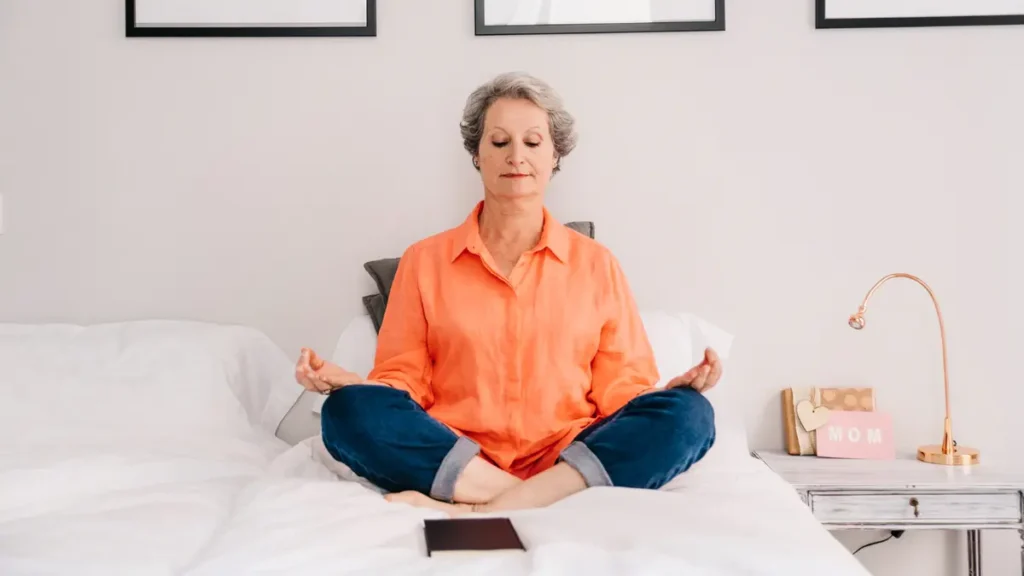Senior woman sitting cross-legged on bed in meditation pose, practicing morning breathing exercise with eyes closed