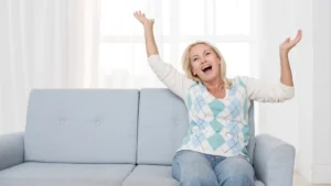 Cheerful middle-aged woman sitting on sofa with arms raised and laughing joyfully in a bright living room, radiating morning energy and positivity.