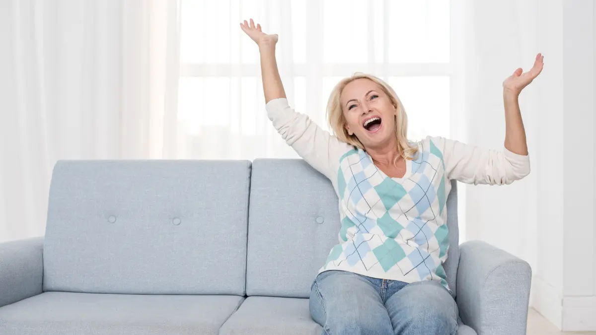 Cheerful middle-aged woman sitting on sofa with arms raised and laughing joyfully in a bright living room, radiating morning energy and positivity.