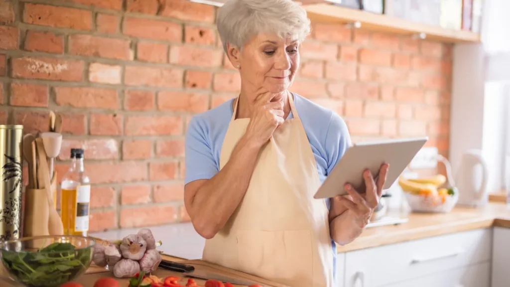 Mature woman wearing an apron looks at a digital tablet to log dietary metrics next to fresh tomatoes and peppers on a kitchen counter.