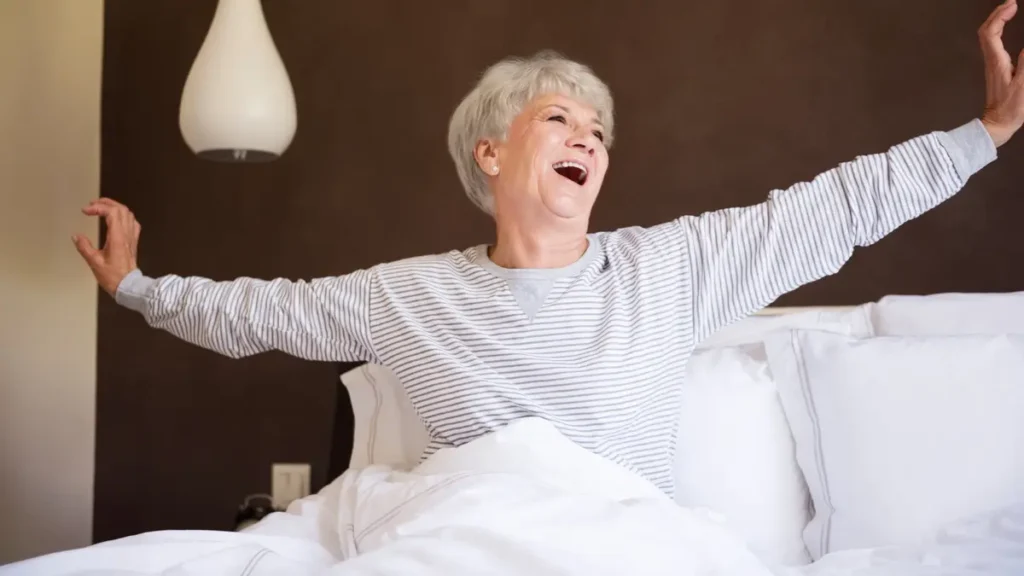 Senior woman stretching arms wide in bed after waking up, smiling joyfully in the morning.