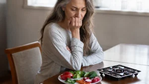 Older woman looking concerned while choosing between healthy vegetables and chocolates, highlighting hidden diet mistakes that weaken bones.