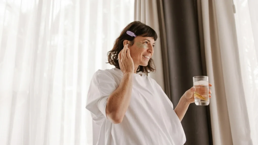 Middle-aged woman in white shirt holding glass of lemon water near bright window with natural morning light, smiling during wellness routine.