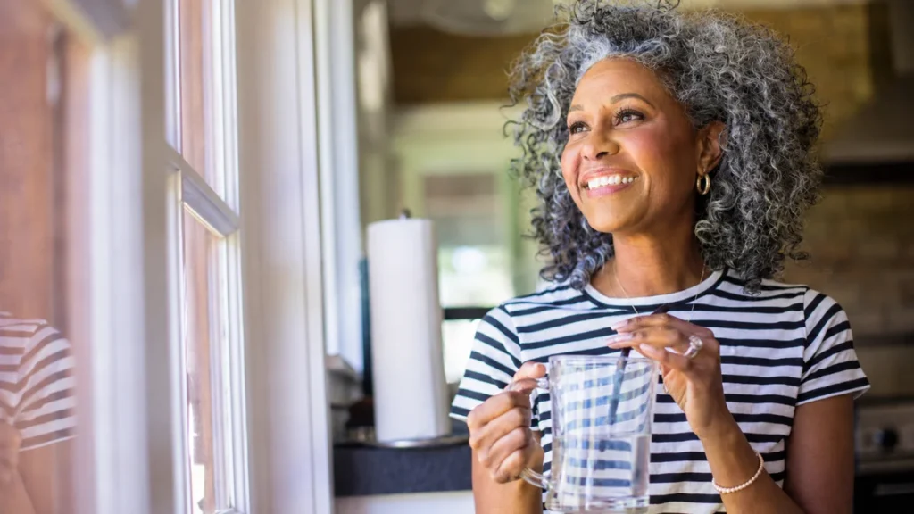 Black woman in her late 50s holding a glass mug and smiling toward a sunlit window, radiating the lifted mood that follows weeks of cutting sugar.