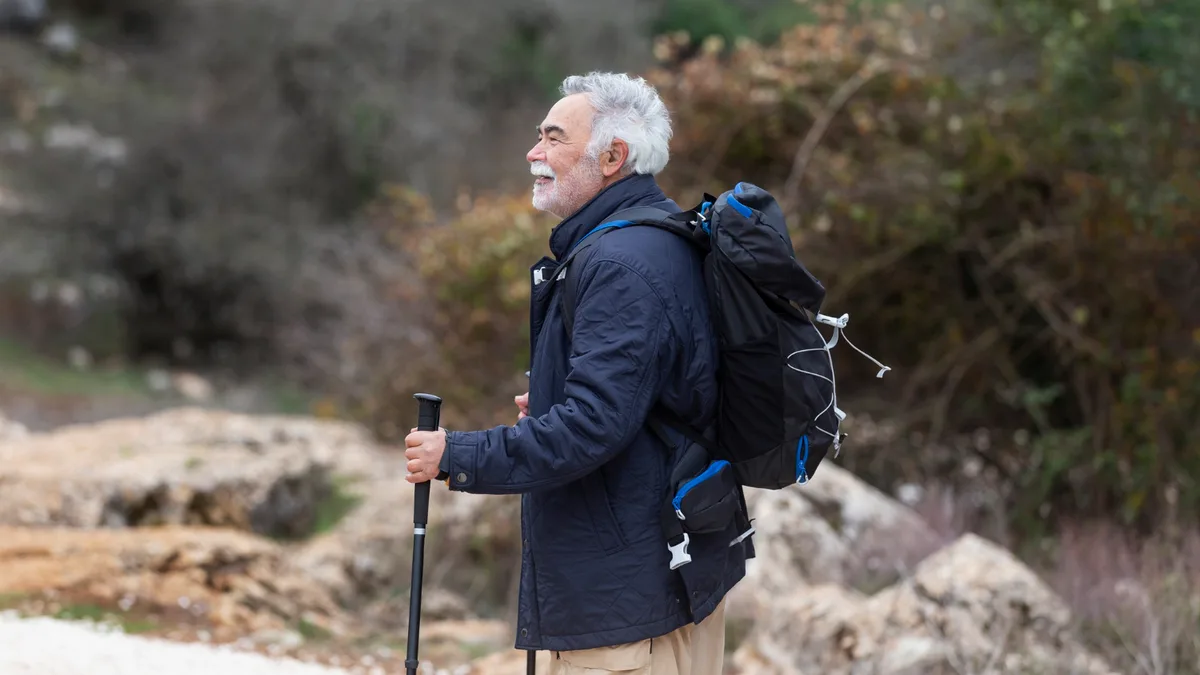 Senior man rucking outdoors with weighted backpack and trekking pole on rocky trail, smiling and enjoying low impact exercise.