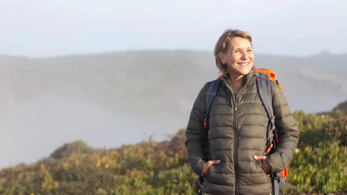 Senior woman rucking outdoors with a backpack smiling confidently in morning light, representing energy and vitality after 50.