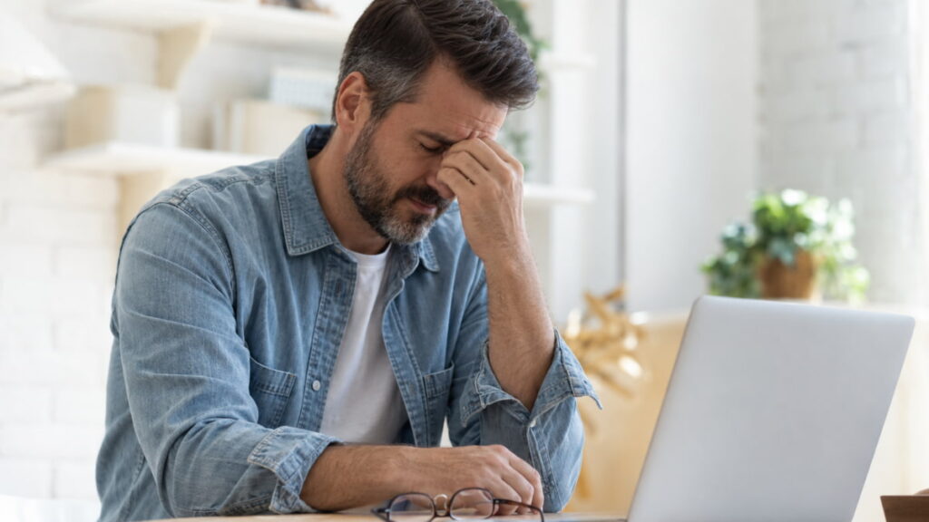 Man sitting at desk with hand pressed to forehead and eyes closed reflecting nervous system freeze and mental exhaustion.