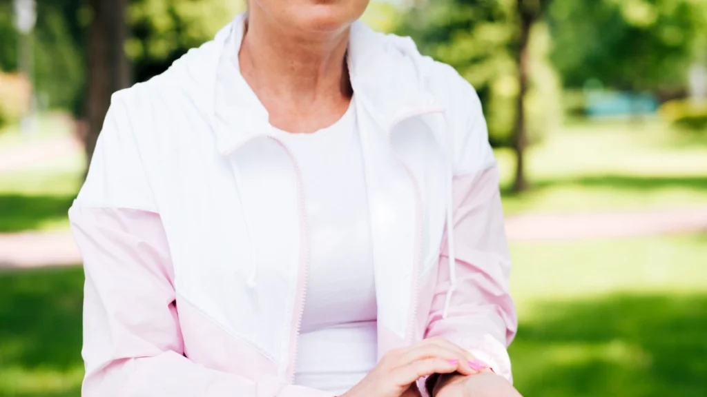 A mature woman in her 60s pauses in a park to check her pulse on a smartwatch, monitoring her heart rate