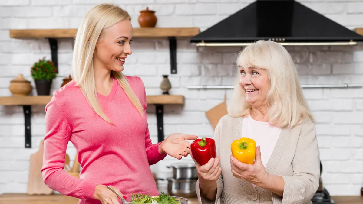 Mature woman smiling while holding a red bell pepper, talking with a younger woman in a bright kitchen as they prepare a healthy meal together.