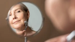 Mature woman using a silver facial massage roller while looking in a mirror, focusing on skincare for aging and glycation.