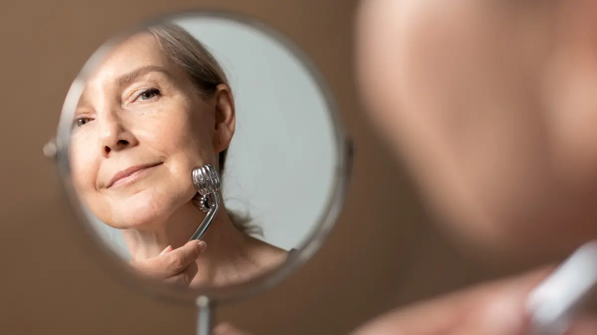 Mature woman using a silver facial massage roller while looking in a mirror, focusing on skincare for aging and glycation.