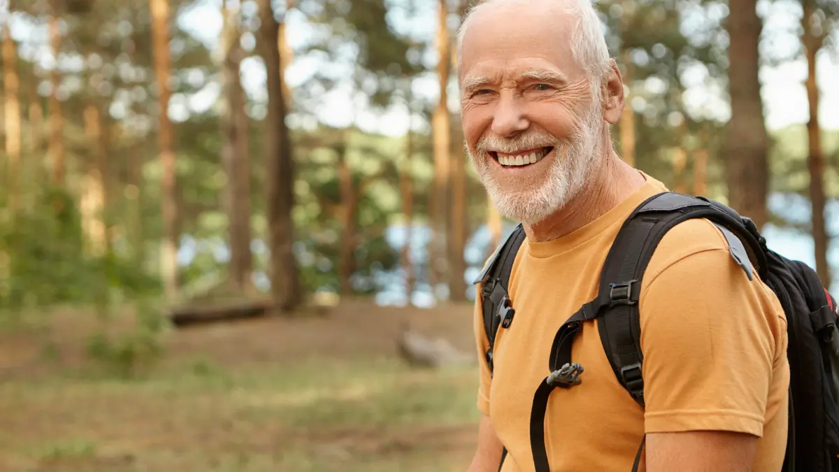 Smiling older man hiking outdoors with a backpack, representing active aging and longevity after 50.