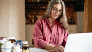 Senior woman researching liver capsules vs real liver on laptop surrounded by supplement bottles to find best nutrition choice.
