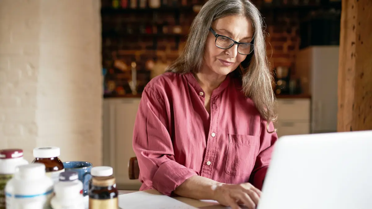 Senior woman researching liver capsules vs real liver on laptop surrounded by supplement bottles to find best nutrition choice.