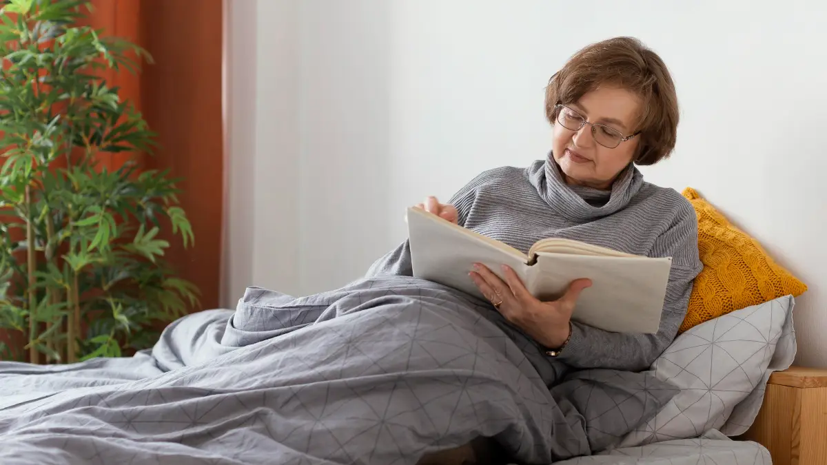 Woman in her 50s lies on lounge chair under thick grey weighted blanket reading book, resting peacefully at home.