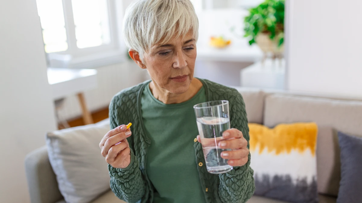 A woman holds a DAO enzyme capsule and a glass of water while sitting on a sofa in a sunlit living room
