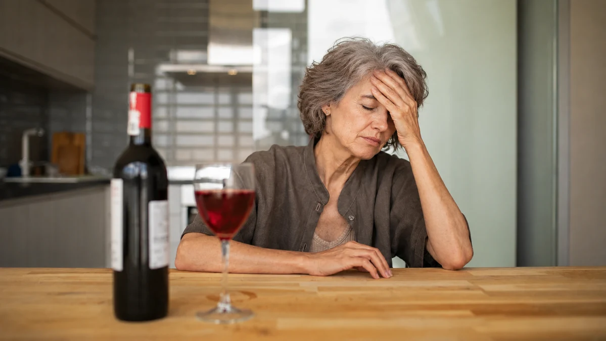 A woman sitting at a table looking unwell next to a glass of red wine showing how alcohol worsens perimenopause symptoms.