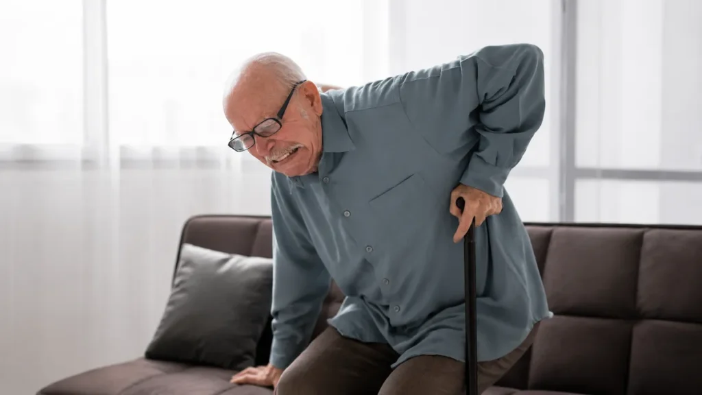 Elderly man with glasses grimacing in pain as he struggles to stand up from a sofa using a walking cane, showing lower back discomfort.