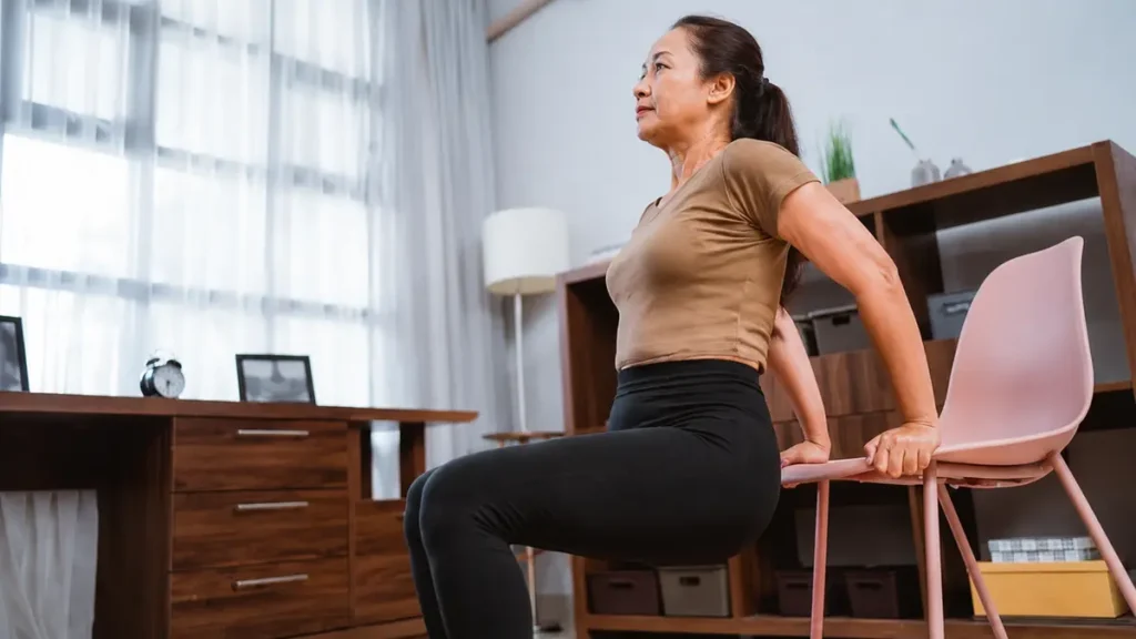 Fit older Asian woman sitting upright on a pink chair in a well-lit home room, holding the armrest while doing seated chair exercises.