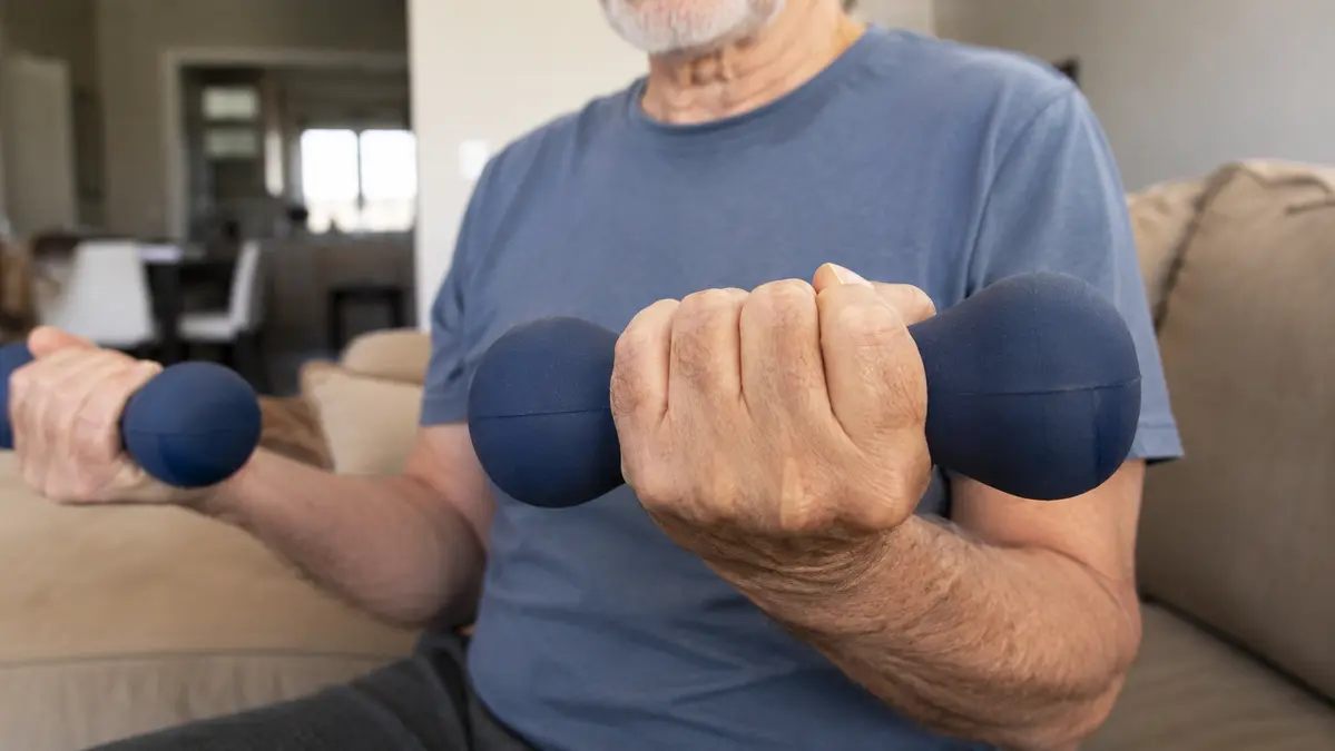 Older man sitting at home on a couch, holding soft neoprene dumbbells in both hands for a gentle exercise.