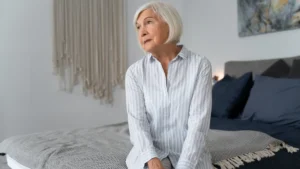 Iron deficiency fatigue in woman after 8 hours of sleep, sitting exhausted on bed with bedroom lights on.