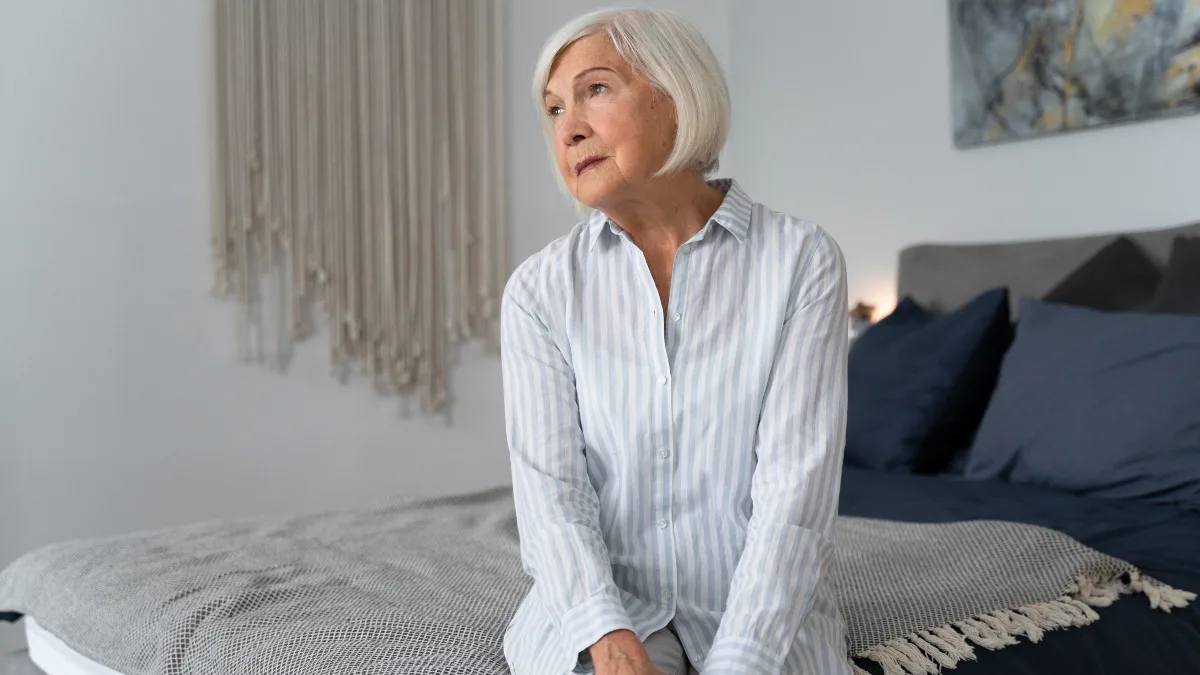 Iron deficiency fatigue in woman after 8 hours of sleep, sitting exhausted on bed with bedroom lights on.