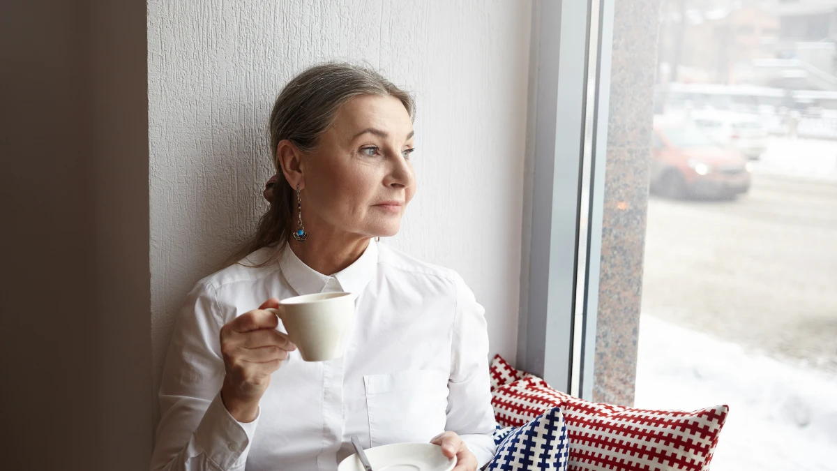 Mature woman sitting by a sunlit window takes a mindful breathing break with a ceramic mug to lower cortisol.