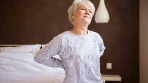 An older woman with short white hair sits on the edge of a bed, clutching her lower back with a pained expression, illustrating the discomfort of morning stiffness
