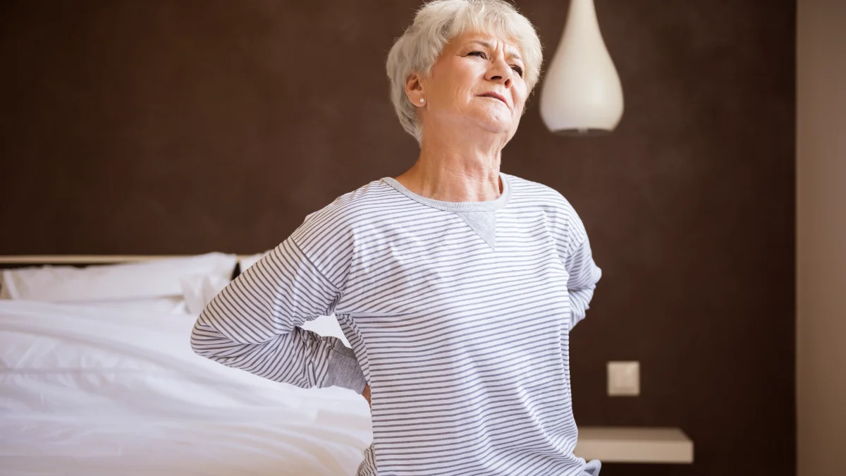 An older woman with short white hair sits on the edge of a bed, clutching her lower back with a pained expression, illustrating the discomfort of morning stiffness