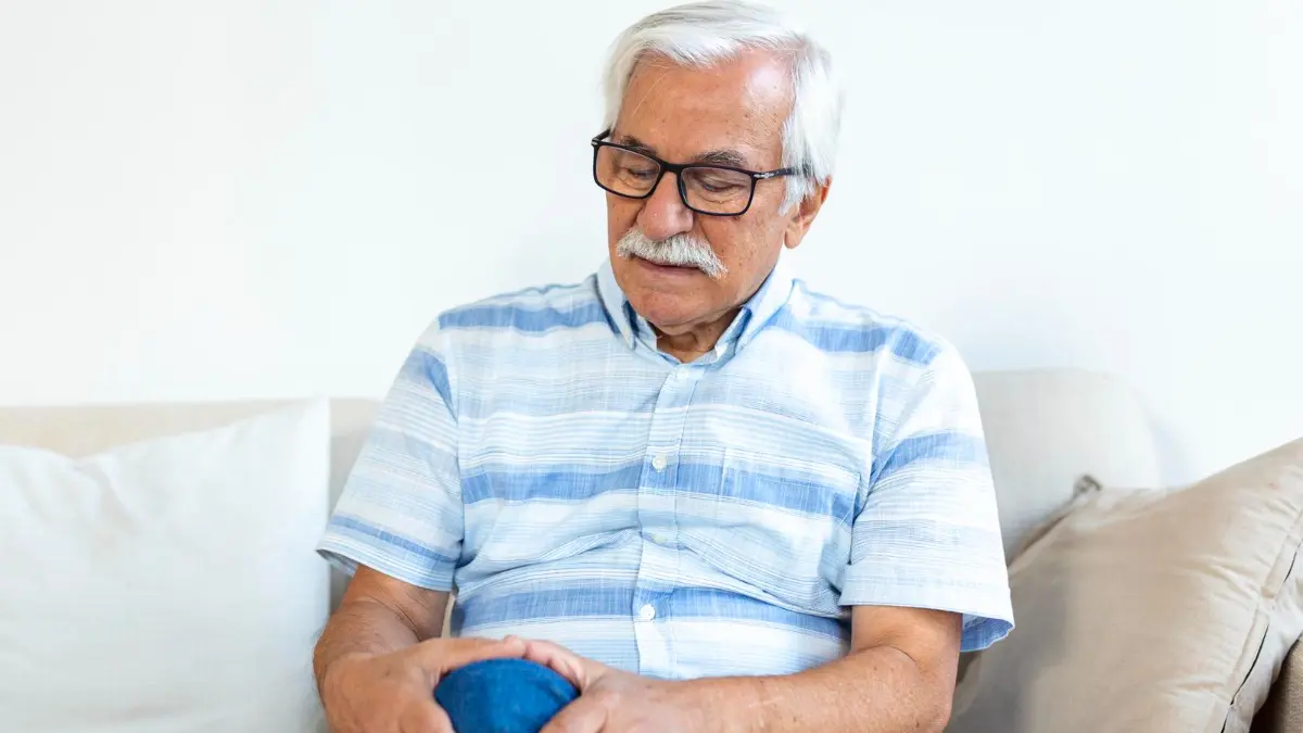 Senior man clutching his throbbing knee on a sofa to identify nightshade-triggered joint pain and inflammation after 50