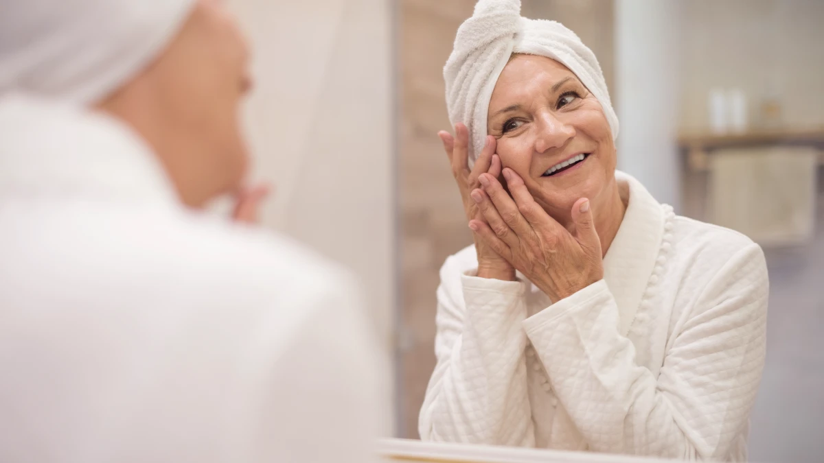 A mature woman examines her skin reflection in a bathroom mirror, contemplating her biological age and cellular health