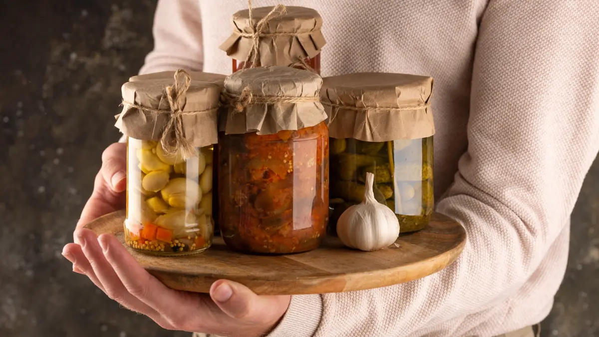 Person holding a wooden tray of assorted glass jars with fermented garlic and pickles to inspect for brine clarity and food safety.