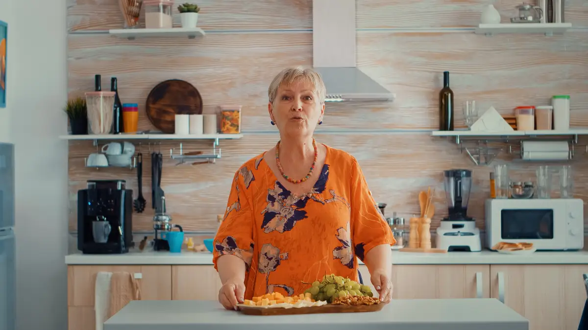 A mature woman in a sunlit kitchen holds a wooden tray with grapes, melon cubes, and walnuts while speaking