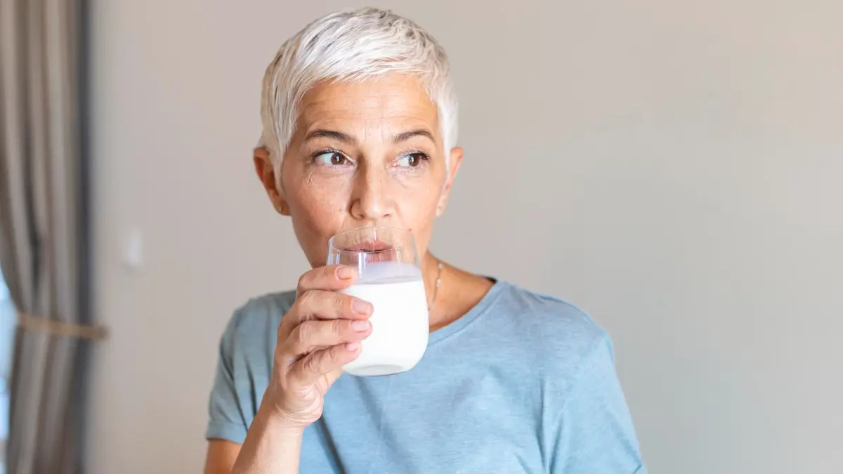  Mature woman with silver hair drinks a glass of unsweetened kefir to prevent the gas and sugar-induced bloating caused by flavored probiotics.