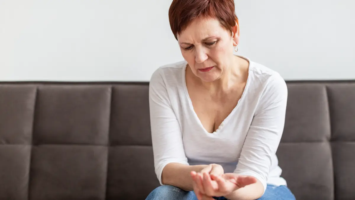 A woman in her 50s rubs her hands while experiencing joint discomfort in a comfortable living room