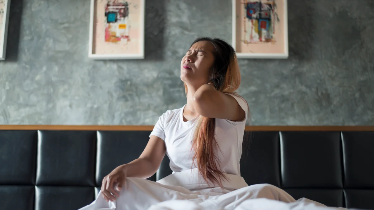 Young woman rubbing her sore neck while sitting in bed in the morning, linked to why you wake up feeling stiff.
