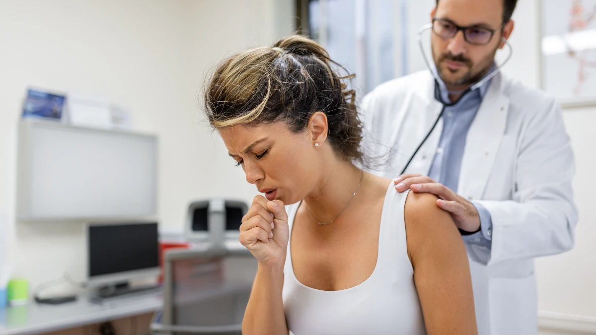 Young woman coughing in a bright clinic as doctor listens with stethoscope to address why lungs feel tight.