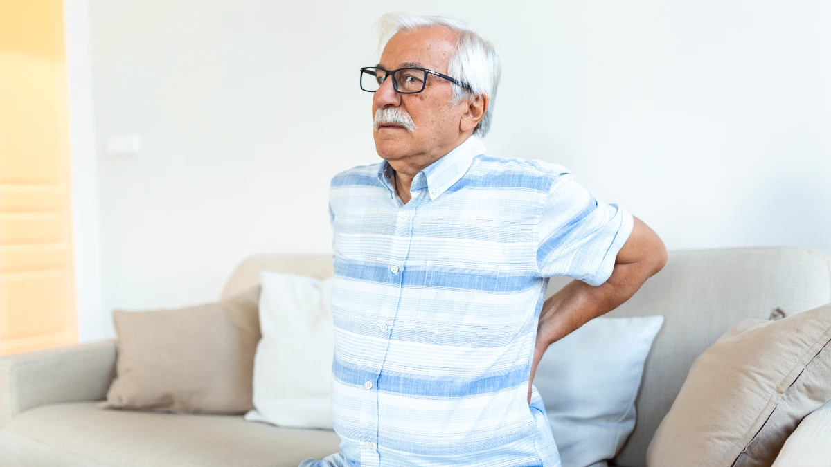 Middle-aged man slightly grimacing and holding his lower back in pain at home, a common symptom of kidney stones or flank pain after 50