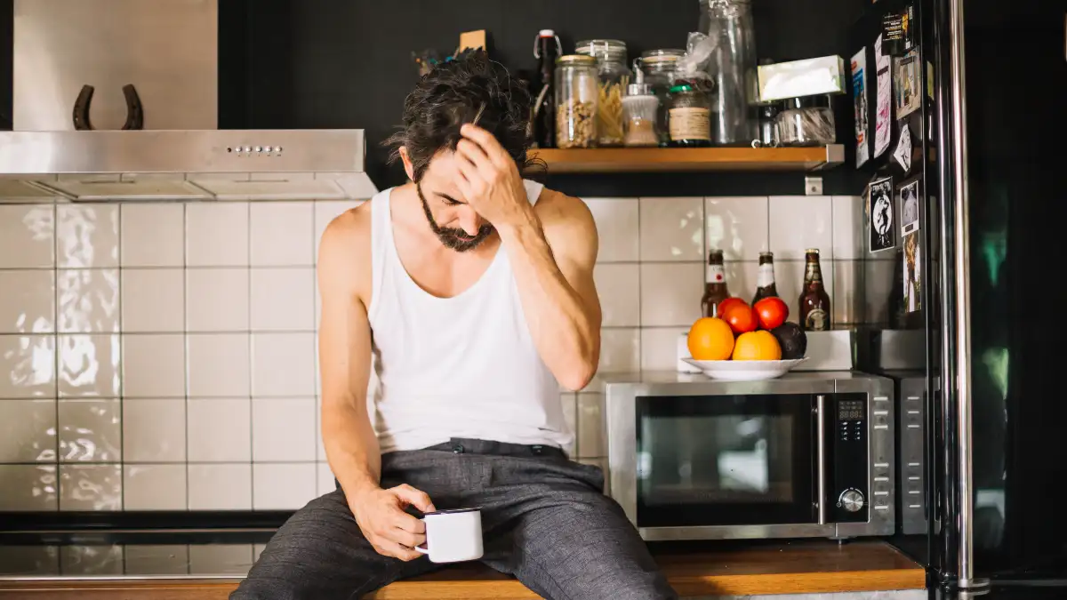 Man in his 50s at kitchen counter holding tea cup pressing two fingers to temple eyes closed leaning forward with sleep inertia grogginess.