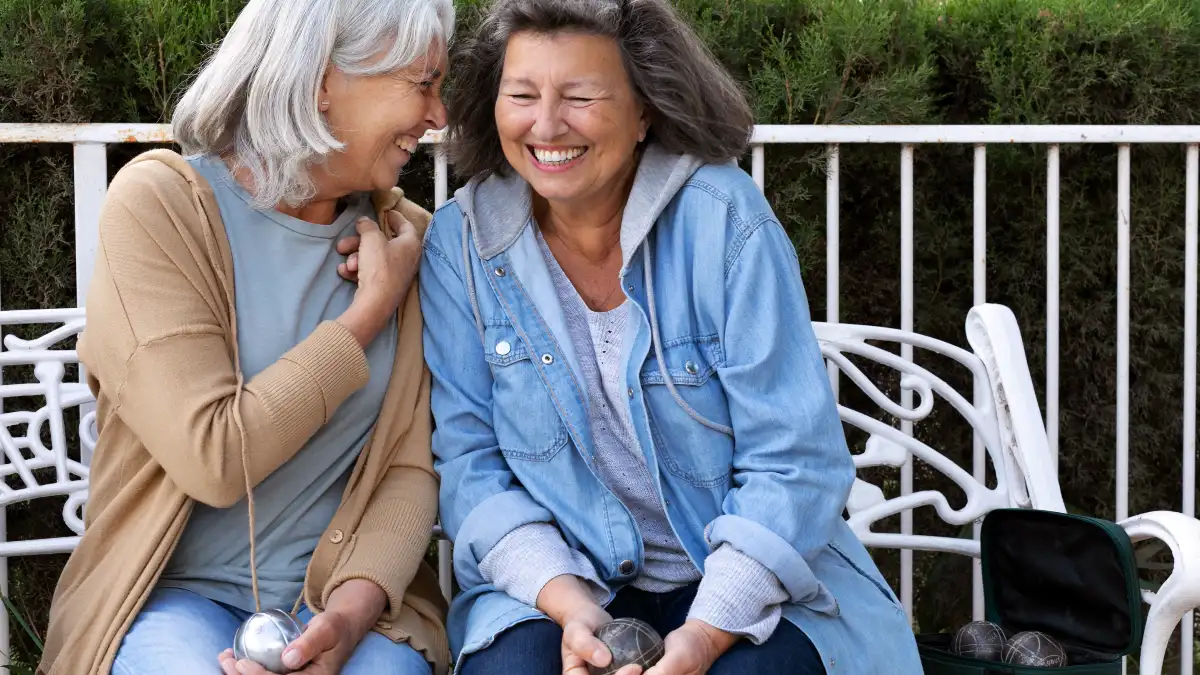 Two women in their 60s share a belly laugh while sitting together on a sunny outdoor patio. 
