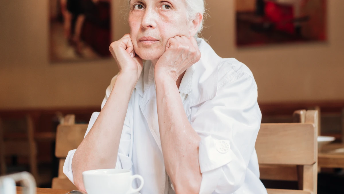  Senior woman looking stressed while sitting in a noisy café, illustrating listening fatigue and hearing strain.