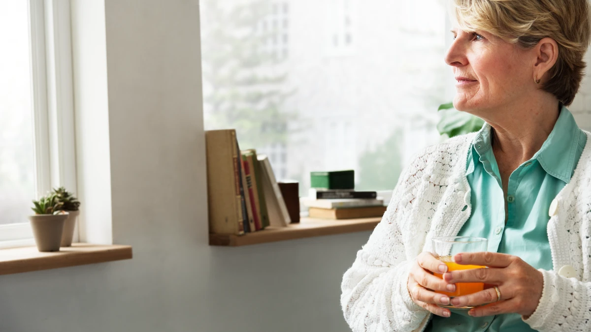 older woman sitting at home holding an orange smoothie, looking thoughtful about healthy drink choices after 50