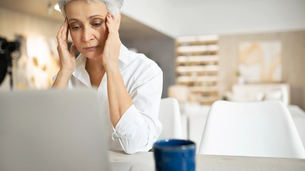 Mature woman sits at a desk with her eyes closed rubbing her temples to manage stress fatigue