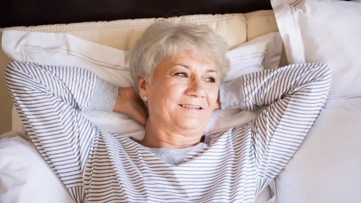 Senior woman in her 60s smiling while lying in cool bedroom looking out open window after deep sleep that triggered brain glymphatic cleaning.