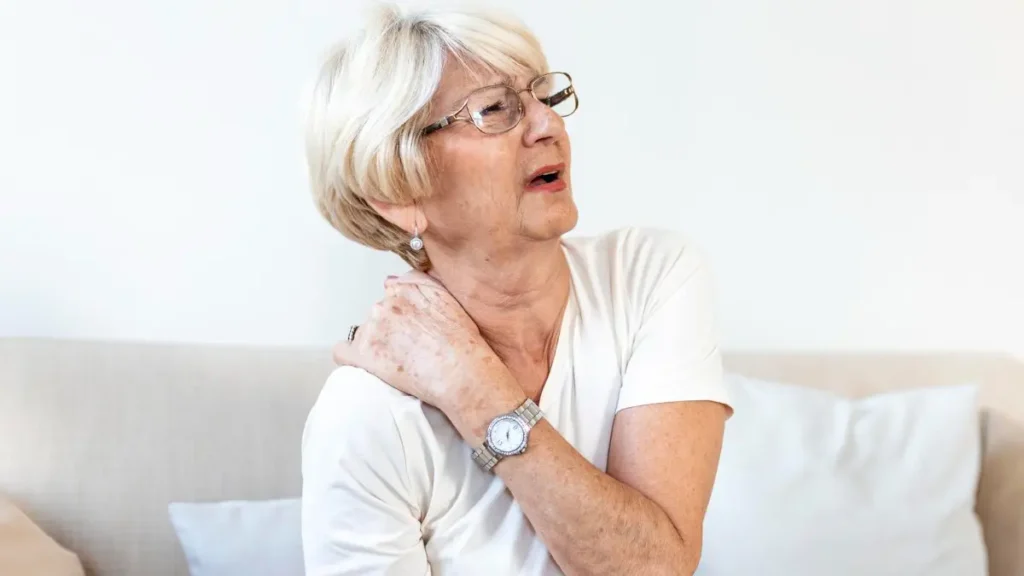 Mature woman in a white shirt sits on a sofa rubbing her stiff shoulder to address inflammaging and cellular waste.