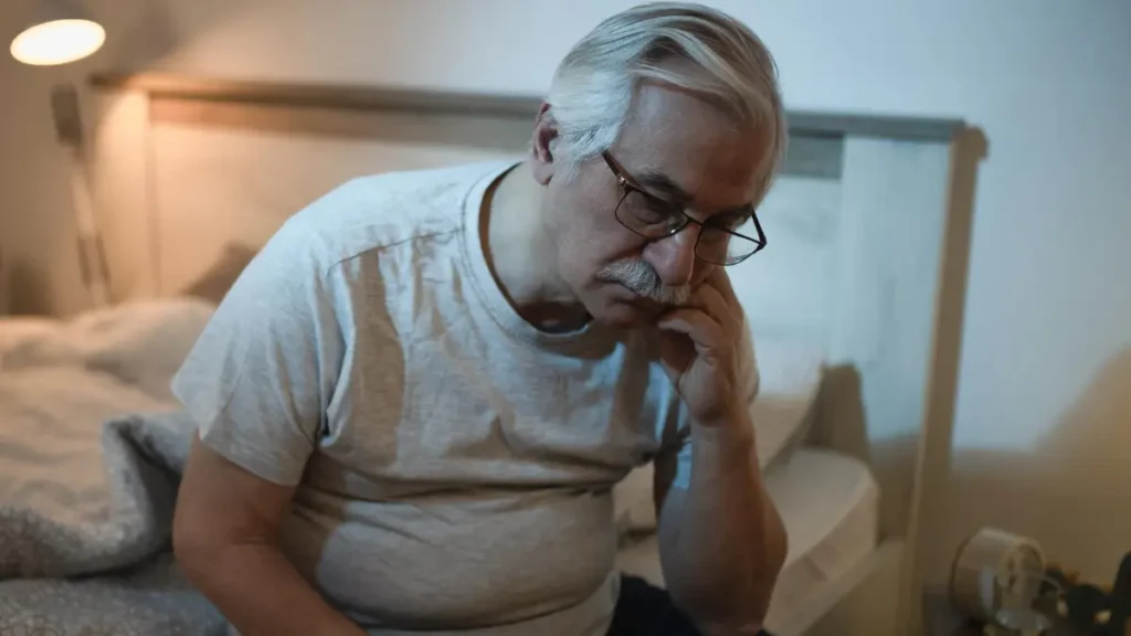A restless mature man sitting on the edge of his bed at night, unable to sleep due to the metabolic heat of a late-night meal.