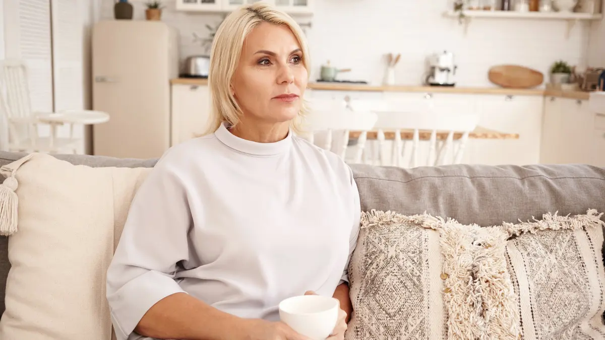 Mature blonde woman on a bright living room couch holding a mug to manage nutrient deficiency memory loss after 50.