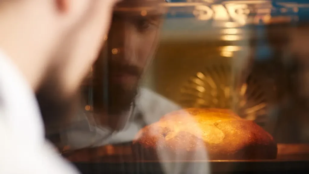 Man closely watching a rustic bread loaf baking inside a steamy home oven without a Dutch oven.
