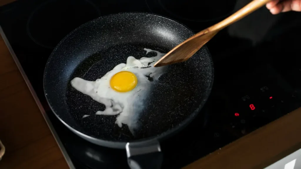 A single egg frying in a dark non-stick pan with a wooden spatula ready to break the yolk.